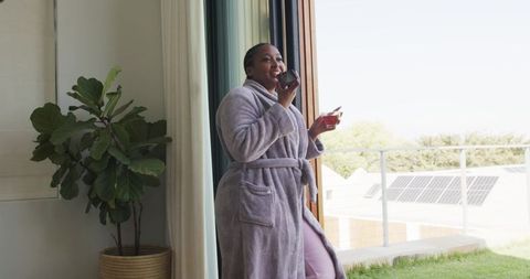 African american woman standing at balcony door wearing grey robe holding mug and glass