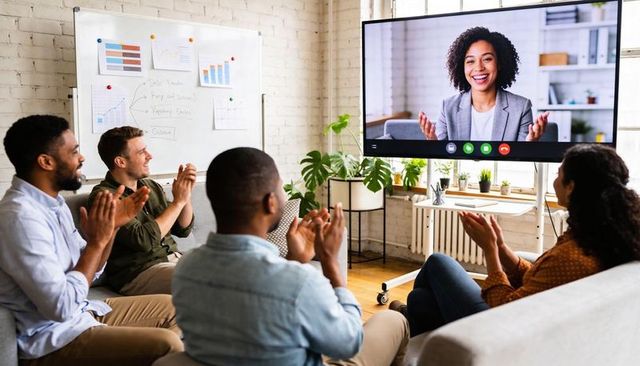 Team applauding remote presenter during virtual meeting in modern office lounge