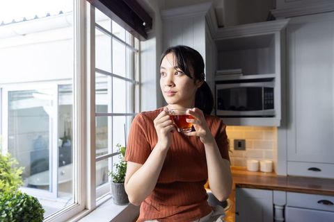 Asian woman sipping tea from glass mug by kitchen window, gazing outside, modern cozy morning