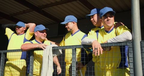 Sunlit baseball teammates leaning on dugout railing wearing yellow jerseys and blue caps