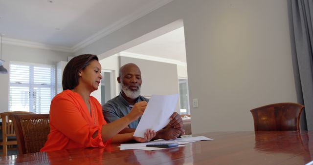 Mature couple discussing and reviewing financial documents while sitting at dining table in modern home. Woman pointing to document as man looks and listens attentively. Great for topics related to personal finance, budgeting, financial planning, teamwork in relationships, domestic life, and home organization.