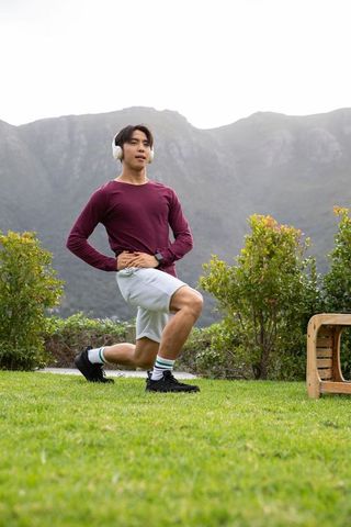 Asian man performing forward lunge with headphones on grassy lawn near mountains