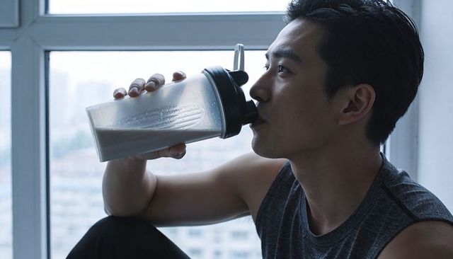 Asian man drinking protein shake by high-rise window during quiet urban workout recovery