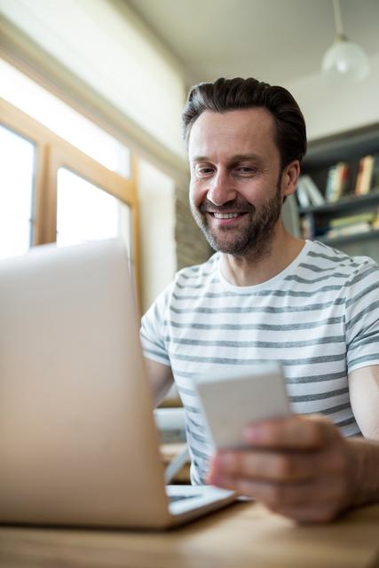 Man smiling while using laptop and mobile phone in coffee shop. Ideal for illustrating remote work, freelance lifestyle, digital communication, and modern technology use in casual settings.