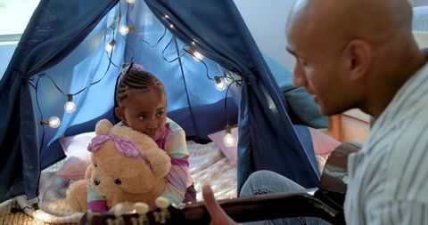 Father playing guitar while african american daughter hugging teddy in cozy indoor teepee