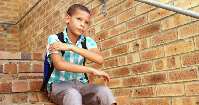 Young boy in plaid shirt sitting on stairs next to brick wall wearing a backpack and looking thoughtful. This image can be used for educational content, back-to-school promotions, or advertisements aimed at children and education. It is a great fit for articles related to school life, childhood development, and outdoor activities.
