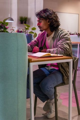 Indian man working on laptop in modern coworking space with notebook, plants, ambient lighting