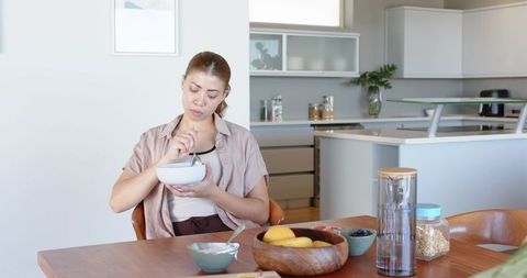 Woman sitting at wooden table holding bowl and stirring breakfast in open-plan kitchen