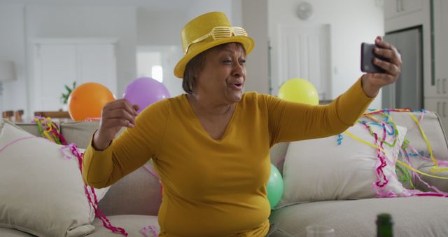 Senior woman wearing bright yellow hat celebrating virtual party at home with colorful balloons in background. She is happily taking a selfie or participating in video call, expressing excitement. Perfect for illustrating modern ways of socializing, virtual celebrations, and joyful moments at home.