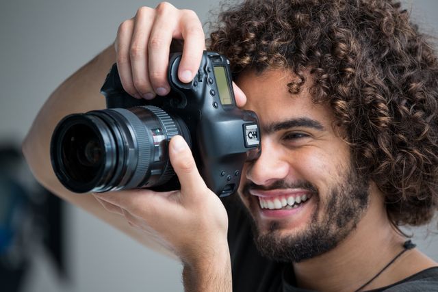 Young male photographer with curly hair smiling while using a digital camera in a studio. Ideal for use in articles or advertisements related to photography, creative professions, hobbies, or lifestyle. Suitable for illustrating concepts of passion, creativity, and professional work environments.