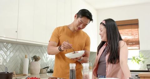 Asian couple cooking together in sunlit modern kitchen, man stirring salad, woman smiling