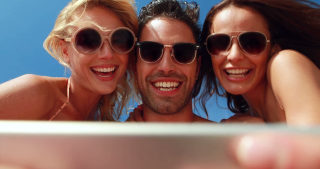 Friends are taking a selfie on a sunny beach, clearly enjoying their time together. They are all wearing sunglasses, indicating a bright sunny day. The image is perfect for websites, marketing materials, travel blogs, vacation packages, and lifestyle promotions as it exudes a sense of fun, happiness, and carefree enjoyment.