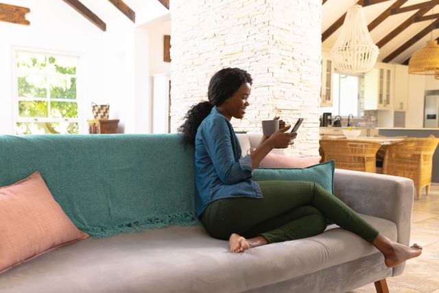 African american young woman having coffee while surfing on smart phone on sofa in living room. Copy space, unaltered, people, lifestyle, home and technology concept.