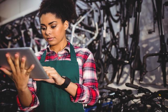Young female bike shop employee using digital tablet, likely managing inventory or customer orders. Ideal for use in themes related to retail, bicycle repair, small business, technology in workplaces, and female empowerment in traditionally male-dominated fields.