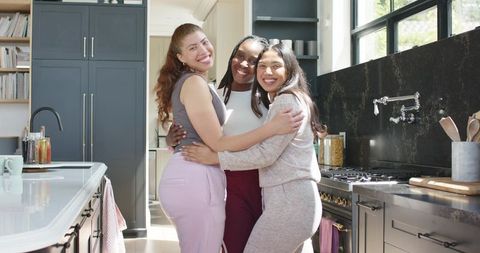Multicultural women hugging and smiling in sunlit modern kitchen embracing friendship