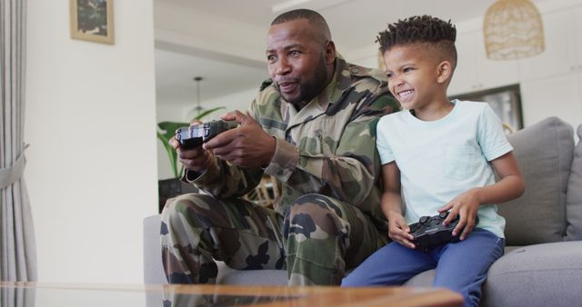 African American father in army uniform enjoying quality time with his young son while playing video games on a couch. Ideal for themes related to family bonding, military families, leisure activities, father-son relationship, and home life.