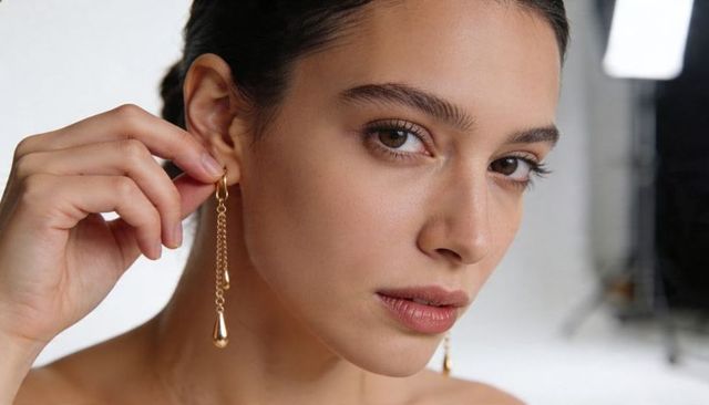 Adjusting gold teardrop earrings closeup portrait bare-shouldered model in studio lighting