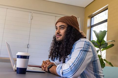Young man working remote with laptop and coffee wearing beanie at bright workspace