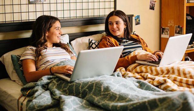 College roommates reclining, studying and chatting in cozy dorm bed with green and yellow blankets