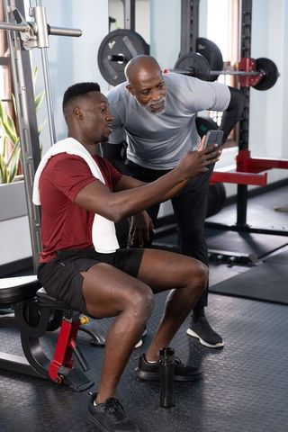 African american senior trainer coaching young athlete checking workout app on bench