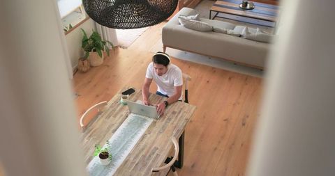 High-angle view man working remotely at wooden dining table in modern living room with laptop and he