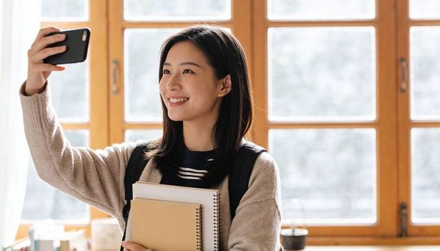Smiling student taking selfie at bright study window holding notebooks and backpack in daylight