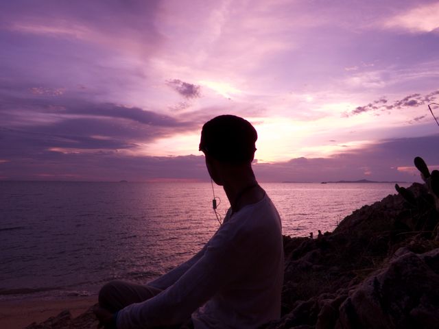 Male preserving tranquility while enjoying peaceful sunset on rocky beachfront. Ideal for themes of relaxation, peace, solitude in nature, and escape from daily stress. Suitable for blogs about mental health, travel, and self-reflection imagery.