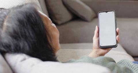 Senior chinese woman reclining on sofa holding smartphone with blank screen mockup