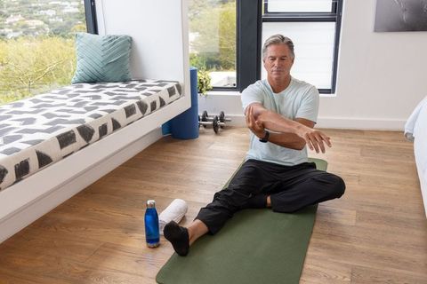Mature man stretching on yoga mat in bright bedroom with dumbbells and foam roller