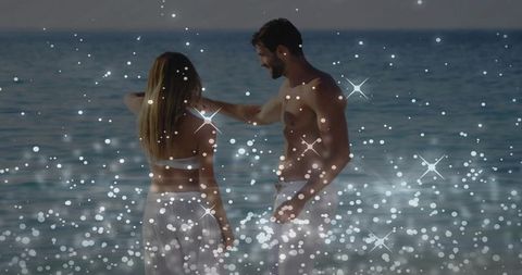 Romantic couple standing in shallow sea at dusk reaching toward shoulder with glowing bokeh