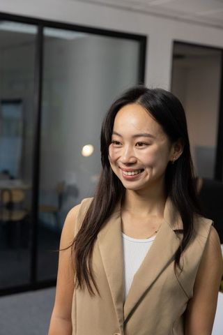 Smiling asian businesswoman standing in modern glass office wearing beige blazer and white top