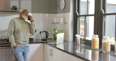 Senior Man in Modern Kitchen on Smartphone Relaxing Near Laptop