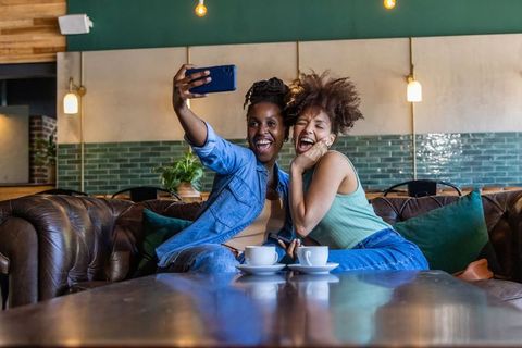 Two african american friends taking selfie in cozy urban cafe lounge, laughing over coffee