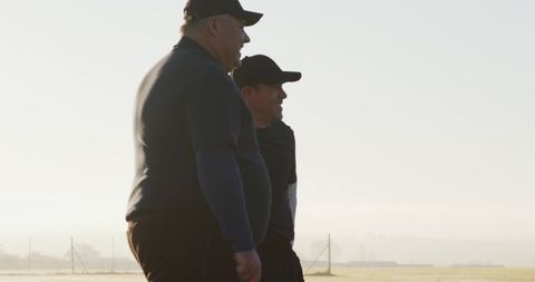 Backlit coaches observing misty sports field at sunrise — two mature men standing