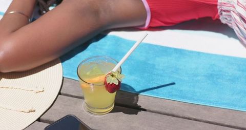 Woman relaxing on poolside towel with citrus cocktail and straw hat, summer leisure