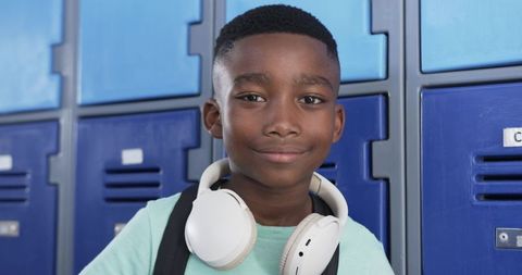 Confident schoolboy wearing white headphones and backpack standing by blue lockers smiling