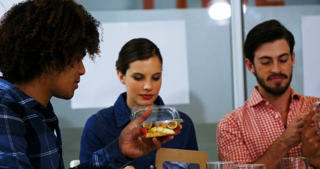 Group of diverse coworkers sharing a healthy lunch in a modern office environment. Two men and one woman are involved in conversation and enjoying their food. Perfect for use in articles about workplace culture, healthy eating habits, modern offices, or corporate teamwork imagery.