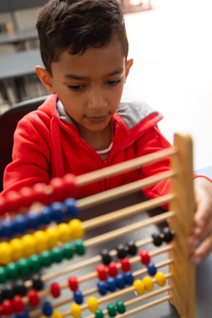 Young biracial boy in red hoodie using abacus in classroom, focused on colorful beads, learning math. Ideal for educational content, early childhood learning materials, school brochures, and math-related articles.