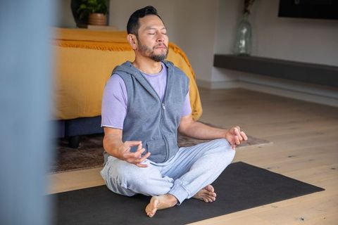 Mature asian man meditating on yoga mat in cozy bedroom for mindfulness and relaxation