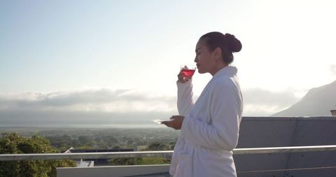 Asian woman in bathrobe enjoying morning tea on balcony overlooking coastal view