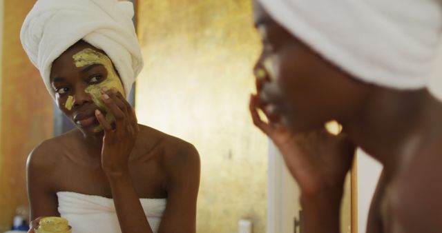 African American woman wrapped in towel applying face mask in bathroom mirror. Ideal for content related to self-care, beauty routines, skincare tutorials, wellness blogs, and personal care advertisements.