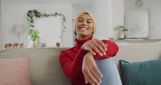 Image of happy biracial woman in hijab sitting on sofa and looking at camera. Lifestyle, spending free time at home concept.