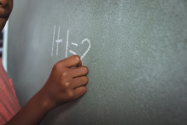 Cropped image showing student writing a simple math equation on a blackboard with chalk. Image highlights educational concept, suitable for use in educational content, school brochures, teaching resources, websites, or articles related to early childhood education, learning process, and classroom activities.