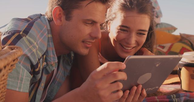 Couple lying on blanket outdoors, enjoying a picnic while watching content on tablet. Suggests themes of leisure, relaxation, and modern technology. Ideal for lifestyle blogs, technology advertisements, or articles about outdoor activities and couples bonding.