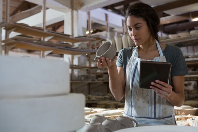 Female potter holding a ceramic cup while using a digital tablet in a pottery workshop. Shelves filled with pottery items are visible in the background. This image can be used for articles on pottery making, artisan crafts, small business promotion, creative processes, and the integration of technology in traditional crafts.