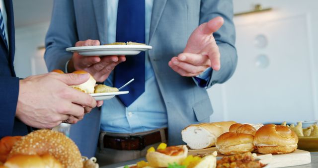 Business professionals in formal attire networking while holding plates during a buffet lunch. Ideal for corporate events, professional networking, office common areas, social interactions in business environments, and casual business meetings.