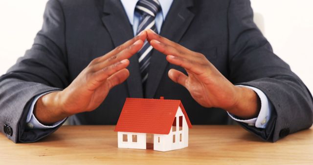 Businessman in suit placing hands over small house model symbolizing protection, security, and real estate investment. This can be used for themes related to property insurance, real estate investment, and home security. Applicable for advertisements or content promoting mortgage services, real estate agencies, insurance policies, and financial planning.