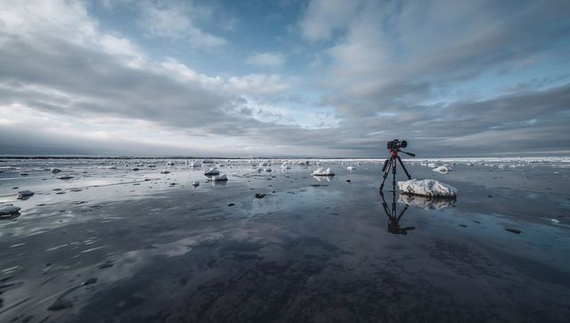 Tripod camera reflecting on icy tidal flat at low tide under dramatic overcast sky