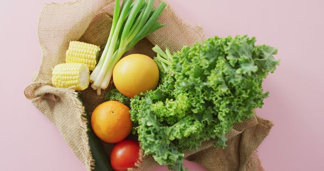 Colorful assortment of fresh vegetables and fruits in a burlap bag. Includes greens, corn, grapefruit, tomato, cucumber, and green onions on a pink backdrop. Ideal for promoting healthy eating, organic food, farm produce, nutrition campaigns, and kitchen decor.