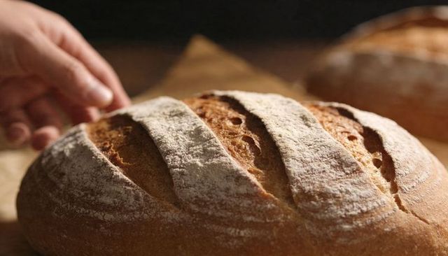 Rustic artisan loaf with crusty scoring and floured hand resting on wooden board, warm close-up